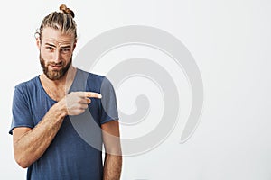 Manly beautiful guy with trendy hairstyle and beard having flirty face expression pointing a side with finger on white