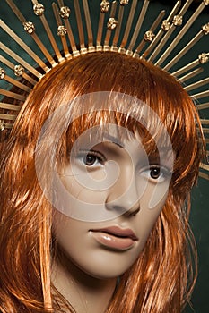Mannequin with bright long red hair wearing a spiky golden crown posing on a green background