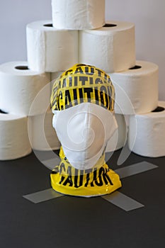 Mannequin head wrapped in yellow caution tape and wearing a mask, with rolls of toilet paper