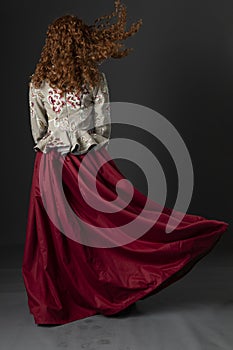 A mannequin with long curly red hair wearing a renaissance-style bodice and red skirt against a studio backdrop