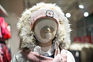 A mannequin on a shop window in clothes: a jacket with a hood, a pink hat with a flower and a pink scarf