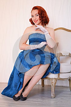Mannered beautiful vintage woman dressed in blue retro dress sitting on a chair alone and smiling on white background