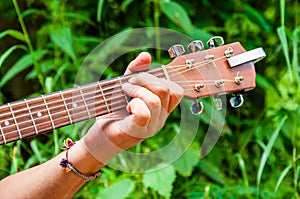 Mans hand holding chord E on a wooden acoustic guitar neck fretboard on scenic green flora background