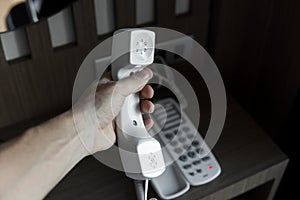 Mans hand holding handset of white vintage telephone with grey battoms on a brown table in hotel.