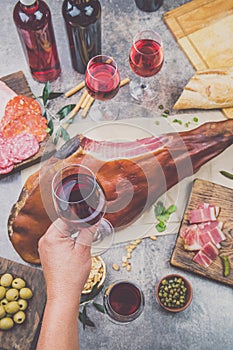 Mans hand holding wine glass frome above table with spanit or italian appetizers, whole leg of ham serrano and red an