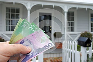 A mans hand holds NZ dollar bills against a front of North American hous