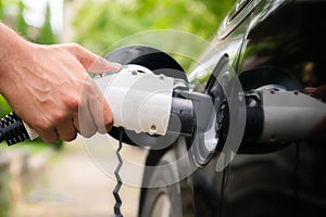 Mans hand inserting charger plug into electric car in green environment background. New energy vehicle, NEV is being loaded with