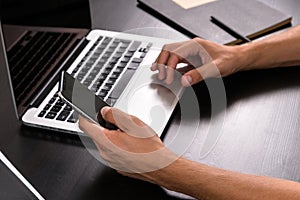 Mans hands typing on smart phone while working on laptop compute