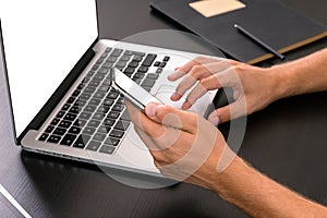 Mans hands typing on smart phone while working on laptop compute