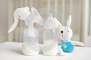 Manual breast pump, bottle with milk for baby and bunny toy over blurred background in children`s room.