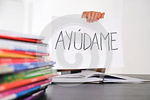 Many books, exercises books on the table and kid hand holding sheet of paper with Spanish word Auydame - Help me -