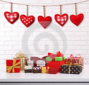 Many colorful gift boxes with ribbon bow on white table and white brick wall with vintage red heart shape garland. Valentines day