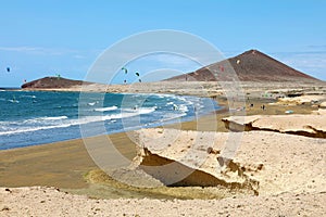 Many colorful kites on beach and kite surfers riding waves and flying during windy day in canarian El Medano in Tenerife with