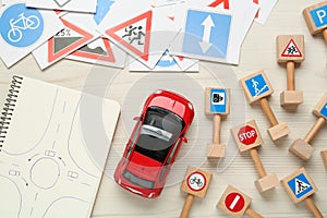 Many different road signs, notebook with sketch of roundabout and toy car on white wooden background, flat lay. Driving school