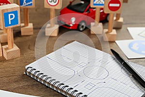 Many different road signs, notebook with sketch of roundabout and toy car on wooden table, closeup. Driving school