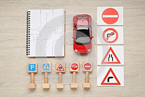 Many different road signs, notebook with sketch of roundabout and toy car on wooden table, flat lay. Driving school