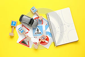 Many different road signs, notebook with sketch of roundabout and toy car on yellow background, flat lay. Driving school