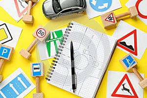 Many different road signs, notebook with sketch of roundabout and toy car on yellow background, flat lay. Driving school