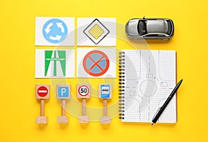 Many different road signs, notebook with sketch of roundabout and toy car on yellow background, flat lay. Driving school