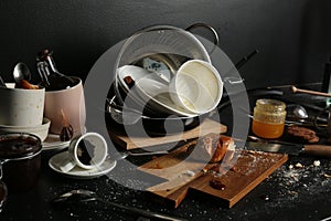 Many dirty utensils, dishware and food leftovers on black countertop. Mess in kitchen