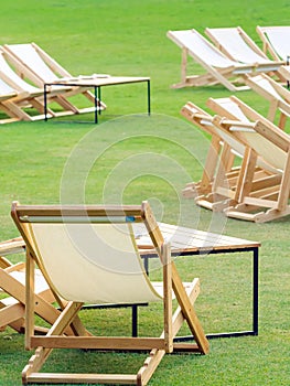 Many empty white deck chairs with tables in lawn is surrounded by shady green grass. Comfortable on outdoor patio chairs in garden