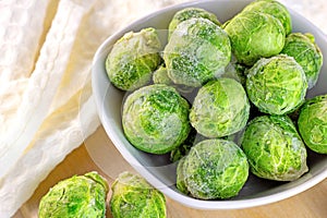 Many frozen whole brussels sprouts in white bowl on light wooden background.