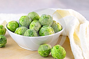 Many frozen whole brussels sprouts in white bowl on light wooden background.