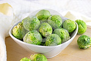 Many frozen whole brussels sprouts in white bowl on light wooden background.