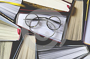 Many multicolored thick open books stand on a dark background. On the books are old round glasses and an open notebook with a