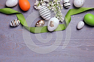 Many painted Easter eggs, branch with lilac flowers and ribbon on grey wooden table, flat lay. Space for text