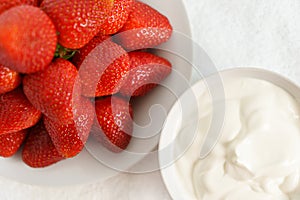 Many red ripe jucy strawberry at white plate close to cup of sour cream on table in light room