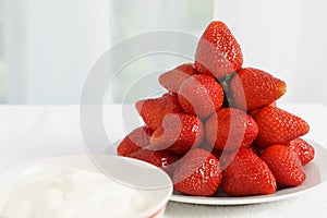 Many red ripe jucy strawberry at white plate close to cup of sour cream on table in light room