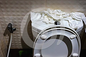 Many of soft toilet paper on top of commode reservoir lid in bathroom. A lot of hygienic paper on flush tank was torn up and