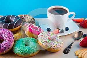 Many tasty donuts with cup of aroma coffee, fruits and one bitten pink doughnut on light stone background.