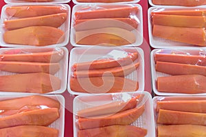 Many tropical papaya slices in styrofoam trays packed with food film on light red table background