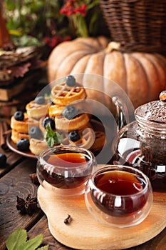 Many waffles are stacked on a plate, decorated with mint leaves and blueberries. Tea in a glass teapot and two small bowls