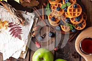 Many waffles are stacked on a plate, decorated with mint leaves and blueberries. Tea in a glass teapot and two small bowls