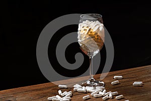 Many white medicine tablets filled in a wine glass. Pills spilled on wooden table. White refillable capsules, pills. Danger.