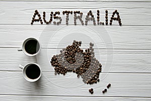 Map of the Australia made of roasted coffee beans laying on white wooden textured background with two coffee cups