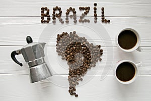 Map of the Brazil made of roasted coffee beans laying on white wooden textured background with two coffe cups and coffee maker