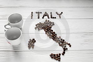 Map of the Italy made of roasted coffee beans laying on white wooden textured background with two cups