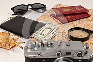map, two passports, a magnifying glass, money in a black leather wallet, an old film camera, sunglasses and a shell on a white wo