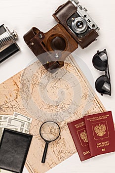 Map, two passports, money in a black leather wallet, an old film camera in a leather case and sunglasses on a white wooden table.
