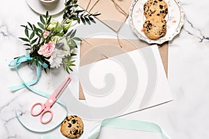 Marble desk with bouquet of flowers, pink scissors, postcard, kraft envelope, cotton branch, oat cookies, invitation card with cop