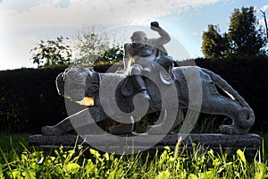 Marble sculpture of chinese tiger and warrior in a garden with green grass and sunset sun as background.