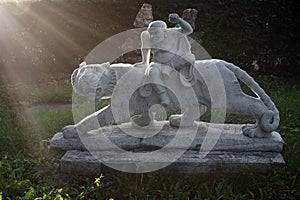 Marble sculpture of chinese tiger and warrior in a garden with green grass and sunset sun as background.