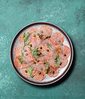 Marbled beef carpaccio with arugula and capers on gray plate, blue background, top view