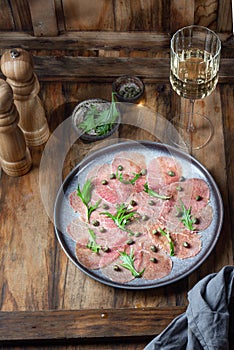Marbled beef carpaccio with arugula and capers on gray plate, with white wine, wooden background, top view