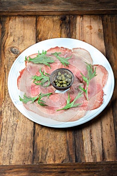 Marbled beef carpaccio with arugula and capers on white plate, wooden background, top view