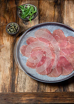 Marbled beef for carpaccio with ingredients arugula and capers on gray plate, wooden background, top view